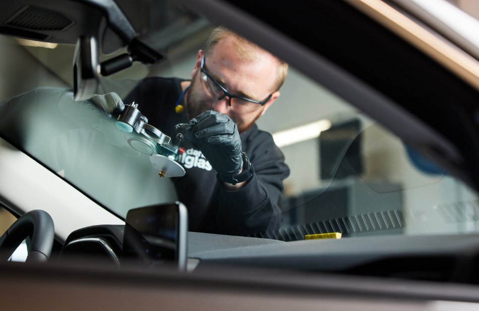 Mand arbejder på bilrude med udstyr, set gennem forruden.